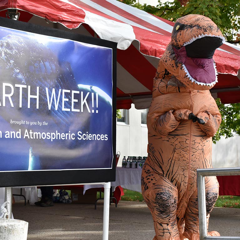 Department mascot, a T. Rex, poses with a sign for "Earth Week"